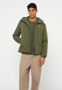 Green padded jacket with a zip front, adjustable hood, and elastic cuffs. Worn over a textured green sweater. Light tan pants.