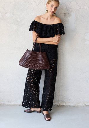 Woman wearing black off-shoulder eyelet jumpsuit, large brown woven handbag, floral earrings, and brown open-toe heels standing against a light wall.