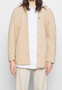 Beige kordjacka med framslutna knappar, två bröstfickor och långa ärmar. Bärs över en vit hoodie, med matchande ljusa byxor.