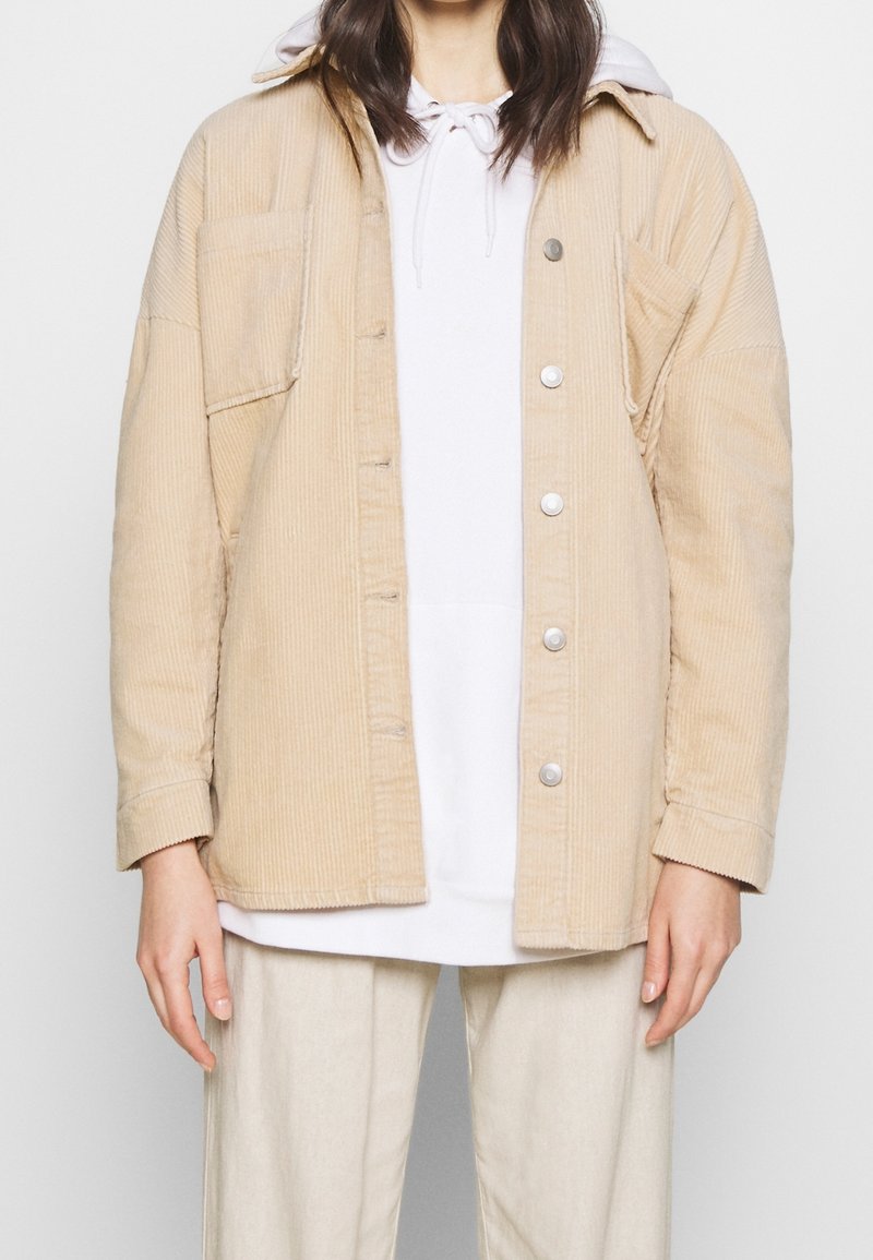 Beige kordjacka med framslutna knappar, två bröstfickor och långa ärmar. Bärs över en vit hoodie, med matchande ljusa byxor.