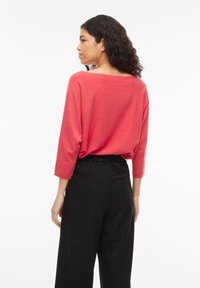 Vrouw met krullend donker haar, gekleed in een losse koraalrode top met lange mouwen en zwarte broek met hoge taille, zijwaarts gekeerd tegen een witte achtergrond.