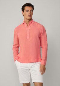 Camicia in lino a maniche lunghe di un vivace corallo con colletto abbottonato e vestibilità leggermente rilassata, abbinata a pantaloni corti bianchi. Tessuto morbido.