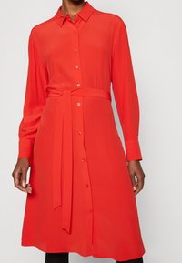 Abito camicia rosso a maniche lunghe realizzato in tessuto morbido, con chiusura a bottoni frontale, vita con cintura e colletto classico.