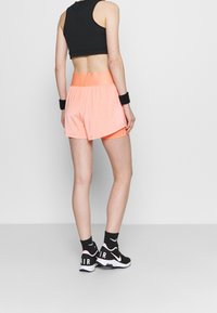 Top corto negro, pantalones cortos deportivos naranjas con forro interior y zapatos negros con detalles rosas. Pulseras texturizadas llevadas en las muñecas.