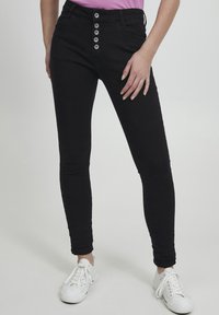Jeans ajustados negros con una cintura alta, cierre frontal de cinco botones y bolsillos laterales. Modelo combinado con zapatillas blancas.