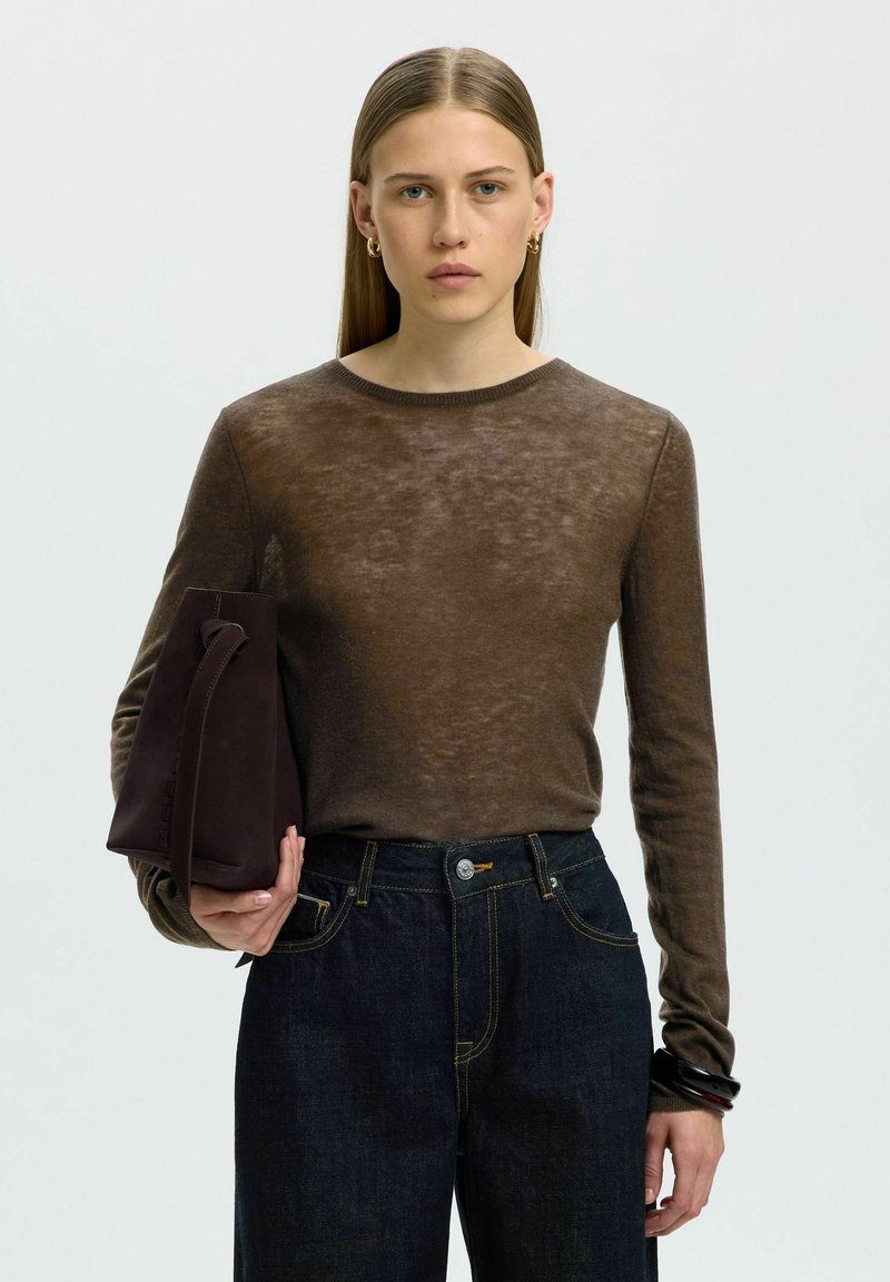 Femme aux cheveux blonds raides porte un haut marron transparent à manches longues, un jean foncé, des boucles d'oreilles créoles dorées, et tient un sac marron foncé plié.