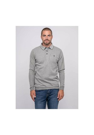 Homme debout face à l'avant, portant un polo gris à manches longues et un jean bleu devant un fond uni.
