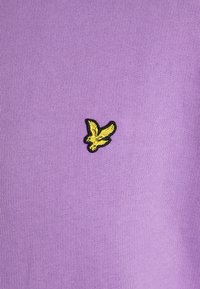 Fioletowa bawełniana koszulka z małym haftowanym logo żółtego ptaka na czarnej obramówce w okolicy klatki piersiowej.