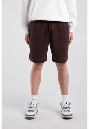 Man draagt donkerbruine korte broek, witte hoodie, witte sokken en witte en grijze sneakers, staand voor een effen achtergrond.