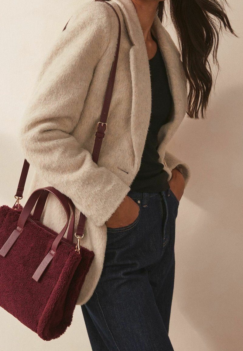Beige fuzzy blazer over a black top, paired with dark jeans. A burgundy textured handbag with short handles rests at the side.