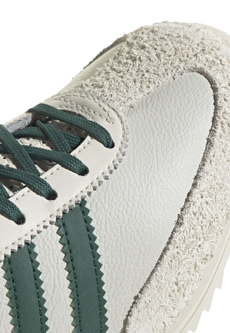 Baskets avec un dessus en cuir blanc texturé, des rayures en zigzag vert et un accent en suède rugueux sur le talon. Des lacets plats verts complètent le design.