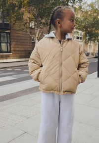Giacca trapuntata beige con chiusura a zip e cappuccio grigio, abbinata a pantaloni della tuta larghi grigi chiari, in piedi su un marciapiede.