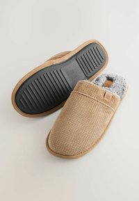 Pantofole lavorate a maglia color sabbia con una parte superiore testurizzata, foderate in morbido grigio e una suola in gomma nera con scanalature orizzontali per la trazione.