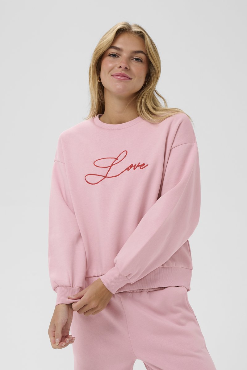 Roza dukserica iz mehkega materiala z dolgimi rokavi in elastičnimi manšetami. Spredaj je okrašena z rdečim vezenim napisom "Love". Ujemajoče hlače dopolnjujejo stilsko podobo.