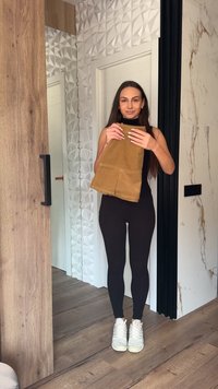 Γυναίκα που στέκεται σε εσωτερικό χώρο κρατώντας ένα διπλωμένο καφέ ένδυμα, φορώντας μαύρο σύνολο και λευκά αθλητικά παπούτσια, με τοιχώματα με υφή και ξύλινο πάτωμα στο φόντο.