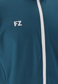 Teal jacka med vit vertikal dragkedja och vit "FZ"-logotyp på bröstet, visar tygtextur och sömnadsdetaljer.