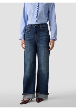 Person wearing wide-leg dark blue jeans with wide rolled cuffs, a light blue striped blouse with ruffles, and maroon pointed heels.