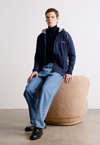 Giacca zip-up blu marina sopra un dolcevita nero, abbinata a jeans larghi azzurri e scarpe nere, seduto su uno sgabello rotondo beige dalla texture particolare.