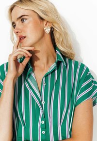 Chemise rayée verte avec des lignes violettes et blanches, à manches courtes et fermeture à boutons. Présente des boucles d'oreilles en forme de cœur en or. Convient pour un usage décontracté.