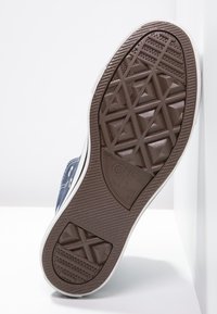 Semelle en caoutchouc marron d'une basket Converse All Star montrant le motif de la bande de roulement et le logo, avec une partie de la chaussure en toile bleue visible.