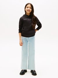 Melnā kapuce ar "TOMMY HILFIGER" logo, zilas plašas bikses un melnas masīvas kurpes. Mati ir gari un taisni, stāvot pret baltu fonu.