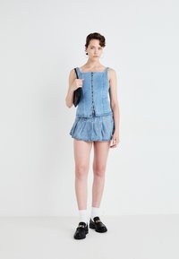 Denim top met knoopdetails en een vierkante hals, gecombineerd met een geplooide denim minirok. Zwarte loafers en een kleine tas maken de look af.