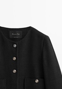 Svart strukturjacka med rund halsringning, silverknappar och två framfickor. Etiketten säger "Massimo Dutti, tillverkad i Rumänien."