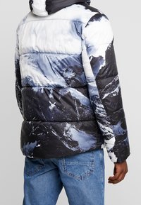 Prošívaná bunda s horským grafikou ve odstínech modré a černé, s lesklým povrchem a kapucí. Nositelná s modrými džínami.