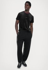 T-shirt de veludo preto com colarinho e logótipo em bordô; combinado com calças de jogging pretas com painéis laterais texturizados e sapatos de atletismo pretos.