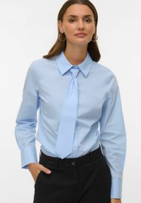 Jasnoniebieska koszula z długim rękawem, zakończona ostrym kołnierzykiem, wraz z dopasowanym krawatem. Wykonana z gładkiego materiału, posiada zapięcie na guziki i luźny krój.