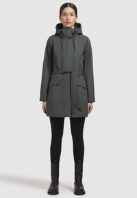 Donkergrijze waterdichte jas met een capuchon, een ceintuur in de taille, twee voorzakken en verstelbare trekkoorden. Een paar zwarte leggings en laarzen.