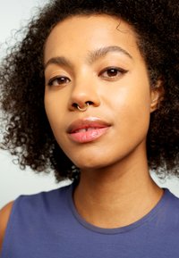 Portrait d'une personne avec des cheveux noirs bouclés, portant un haut sans manches bleu marine, avec un anneau de septum et un maquillage naturel et lisse.