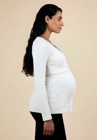 Šviesiai pilkas nėštumo marškinėliai su ilgomis rankovėmis, V formos iškirpte ir prigludusia forma, dėvimi asmens su tamsiais, ilgais, banguotais plaukais.