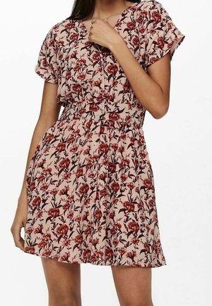 Žena v krátkých růžových šatech na knoflíky s červeným květinovým vzorem a krátkými rukávy, pózující s jednou rukou u klíční kosti.