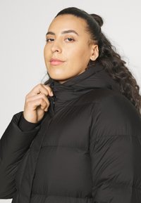 Crna puf jakna s visokim ovratnikom, koja ima ušivene panele i glatku teksturu. Model drži ovratnik jednom rukom.