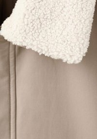 Beige yttertyg med mjukt vitt fleecekrage. Synliga stygn; slät yta på kroppen, i kontrast till den lurviga kragen.