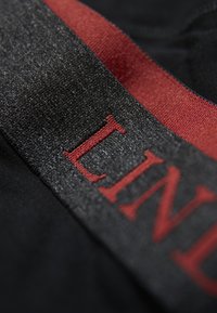 Tessuto nero con una vita testurizzata e lettere rosse che leggono "LIN". Il materiale è liscio con una leggera lucentezza.