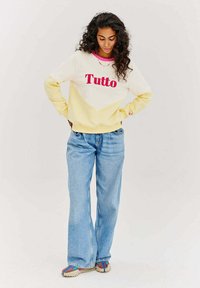 Světle žlutá mikina s červeným textem "Tutto", geometrický design s barevnými bloky; v kombinaci s modrými širokými džíny a multikolorovými vzorovanými botami.