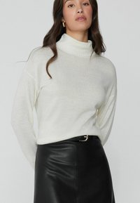 Weißer, gerippter Rollkragenpullover aus weichem Strickmaterial, kombiniert mit einem schwarzen, enganliegenden Lederrock mit subtiler Glanzoptik und Gürtel-Detail.