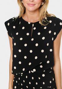 Blouse noire à pois crème, à manches courtes, avec un détail en forme de clé au niveau du col. Fabriquée dans un tissu léger et lisse.