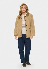 Kviltad beige jacka med brun krage, framfickor och svarta knappar, bärs över en randig tröja och mörka jeans, med en avslappnad passform.