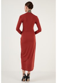 Robe longue à manches longues couleur rouille avec une silhouette ajustée et des fentes sur les côtés, confectionnée dans un tissu lisse et extensible. Des chaussures à talons noirs complètent le look.