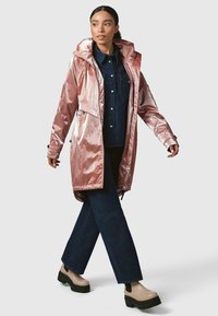 Ilgas rožinės auksinės metalinės lietpaltis su kapišonu, užtrauktukais ir priekinėmis kišenėmis, dėvimas ant džinsinio marškinio ir tamsių džinsų, derinamas su masyviomis smėlio spalvos botais.