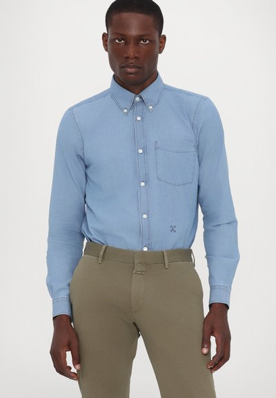 Chemise en denim bleu clair avec col button-down, poche poitrine et boutons sur le devant, associée à un pantalon vert olive. Texture lisse, design ajusté.