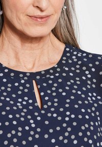 Navyblauwe blouse met een ronde halslijn, voorzien van een sleutelgatuitsnede en een patroon van witte en grijze stippen van verschillende grootte. Lichtgewicht stof.