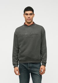 Sudadera gris oscuro con puños y dobladillo acanalados, que presenta un logotipo texturizado "TOMMY JEANS" en el pecho. Lleva puesta una camisa de cuadros rojos.