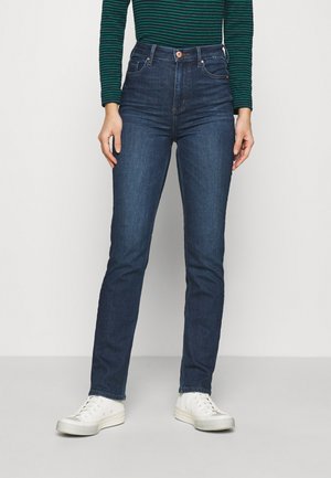 Osoba ubrana w ciemnoniebieskie, przylegające spodnie z wysokim stanem, w połączeniu z białymi trampkami oraz zieloną bluzkę w paski z długim rękawem.