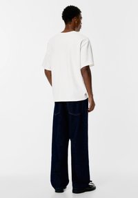 Weißes T-Shirt mit kurzen Ärmeln und lockerer Passform, kombiniert mit dunkelblauen, weit geschnittenen Jeans, die zwei Gesäßtaschen und eine klassische Denimtextur aufweisen.