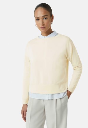 Hellgelbes Sweatshirt mit Rundhalsausschnitt, gerippten Bündchen und einem dezenten Logodetail am Saum, über einem hellblauen Shirt getragen.