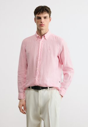 Νέος άνδρας με κοντά καστανά μαλλιά, φορώντας ροζ ανοιχτό πουκάμισο με κουμπιά και λευκό πιετενέ παντελόνι με μαύρη ζώνη, στέκεται μπροστά σε λευκό φόντο.